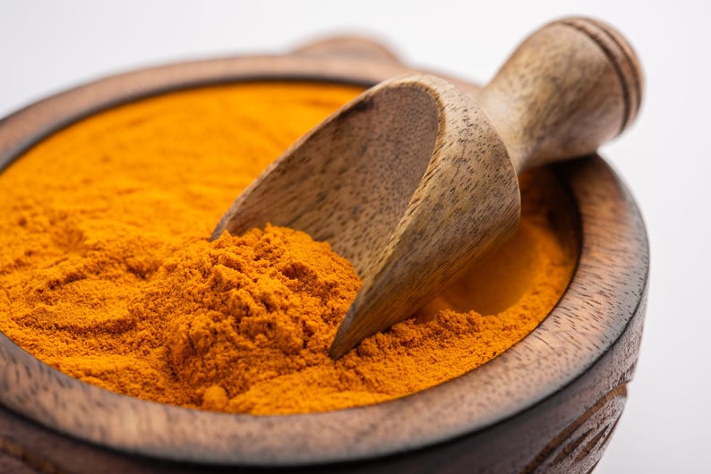 K&ouml;nnte beinhalten: Eine Holzschale gef&uuml;llt mit leuchtend orangefarbenem Kurkuma-Pulver. Eine Holzschaufel ist teilweise in das Pulver eingetaucht.
