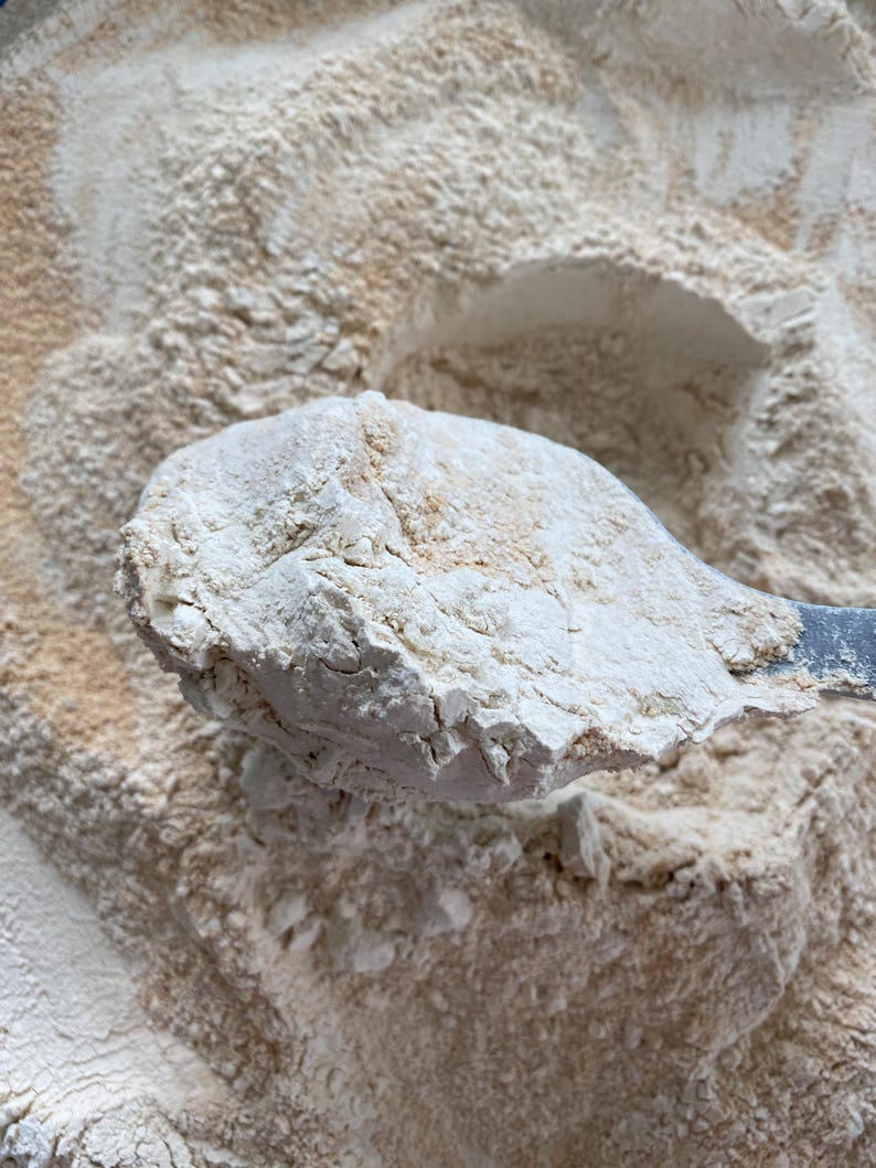 May include: Close-up of a scoop of light brown flour, held by a metal utensil, against a backdrop of more flour. The flour has a slightly coarse texture and a light, natural color, suggesting it may be whole wheat or a similar type.