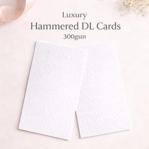May include: Two white, textured DL cards with the text "Luxury Hammered DL Cards 300gsm". The cards are rectangular and have a hammered paper texture. The background is a soft, neutral colour.