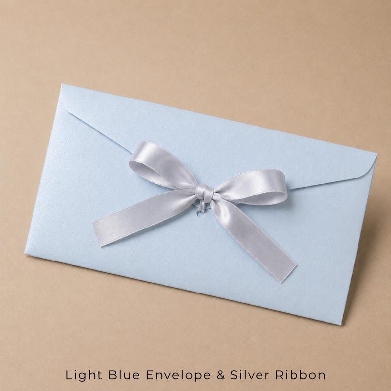 Op de afbeelding: Een lichtblauwe rechthoekige envelop met een gladde, matte afwerking. In het midden van de klep aan de voorzijde bevindt zich een decoratief zilverkleurig satijnen lint dat is gestrikt tot een nette strik. De envelop ligt horizontaal.