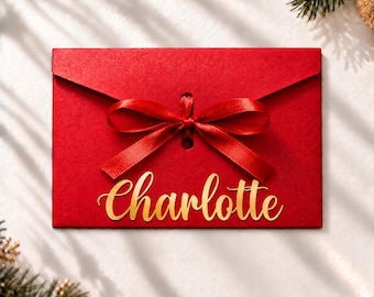 Könnte beinhalten: Ein rechteckiger roter Umschlag mit einer mittig platzierten, zu einer Schleife gebundenen roten Satin-Schleife. Der Name Charlotte ist in goldener Schreibschrift auf dem unteren Teil des Umschlags zu sehen. Er hat eine spitze Klappe.
