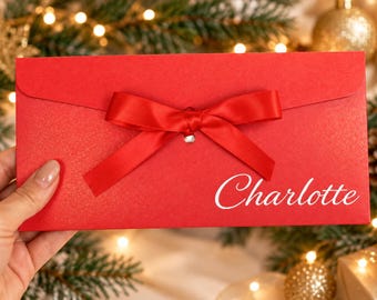 Könnte beinhalten: Ein rechteckiger roter Umschlag mit einer mittigen roten Satinschleife und dem Namen Charlotte in weißer Schreibschrift auf der unteren rechten Seite. Der Umschlag hat einen Klappverschluss mit einem kleinen silberfarbenen Akzent.