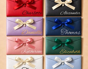 Puede incluir: Ocho sobres rectangulares de varios colores, cada uno con un lazo de satén y un nombre personalizado en letra cursiva. Los colores incluyen rojo, negro, lavanda, azul marino, rosa, verde oscuro, blanco y azul claro con lazos a juego.