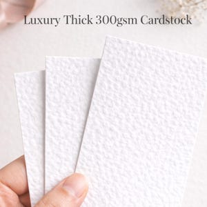 May include: Three white textured cardstock sheets are shown, with the text "Luxury Thick 300gsm Cardstock" at the top. The paper has a subtle, organic texture. The sheets are held by a hand.