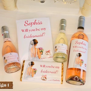 Könnte beinhalten: Ein Brautjungfern-Anfrage-Geschenkset mit einer Flasche Roséwein, einer Flasche Prosecco und einer Schokoladentafel. Die Etiketten auf den Flaschen und der Schokoladentafel lauten "Sophia, Willst du meine Brautjungfer sein?" mit einer Braut-Illustration.