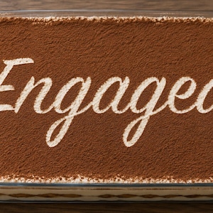 Puede incluir: Un plato rectangular de vidrio lleno de tiramisú, espolvoreado con cacao en polvo. La palabra "Engaged" está escrita en letras blancas en la parte superior, lo que lo convierte en un postre de celebración.