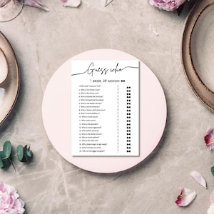 Pode incluir: Um cartão branco com o texto "Guess who" e "Bride or Groom" está em um prato rosa. O cartão tem uma lista de perguntas e gráficos de gravata borboleta. O fundo inclui flores cor-de-rosa e pratos decorativos.