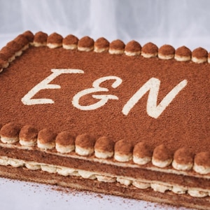 May include: A rectangular tiramisu cake dusted with cocoa powder. The cake is decorated with the letters "E & N" in white icing. The edges are lined with cream-filled ladyfingers, and the layers are visible.