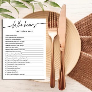 May include: A wedding game card with the text "Who knows THE COUPLE BEST?" is placed on a beige plate with a copper fork and knife. The card has fill-in-the-blank questions. A brown textured napkin is on the right.
