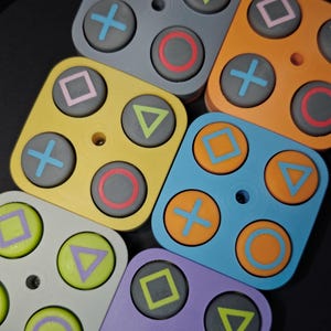 Puede incluir: Una colección de conjuntos de botones de mando de videojuegos cuadrados y coloridos. Cada conjunto presenta cuatro botones con formas icónicas: un círculo, una cruz, un cuadrado y un triángulo. Los botones son de varios colores, incluyendo gris, amarillo, naranja, azul y morado.