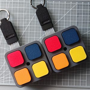 May include: Two keyrings with a grey base and four colourful square buttons: red, blue, yellow, and orange. Each keyring has a black strap and a silver key ring. The items are on a grey grid surface.