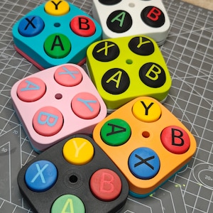 Puede incluir: Una colección de coloridos juguetes fidget cuadrados, cada uno con cuatro botones circulares con las letras A, B, X e Y. Los juguetes son de varios colores, incluyendo azul, rosa, verde, naranja y negro, con colores de botones contrastantes.