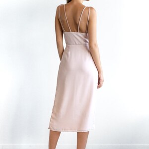 May include: A light pink midi dress with thin spaghetti straps and a simple, elegant design. The dress has a straight silhouette and a slightly longer length in the back.