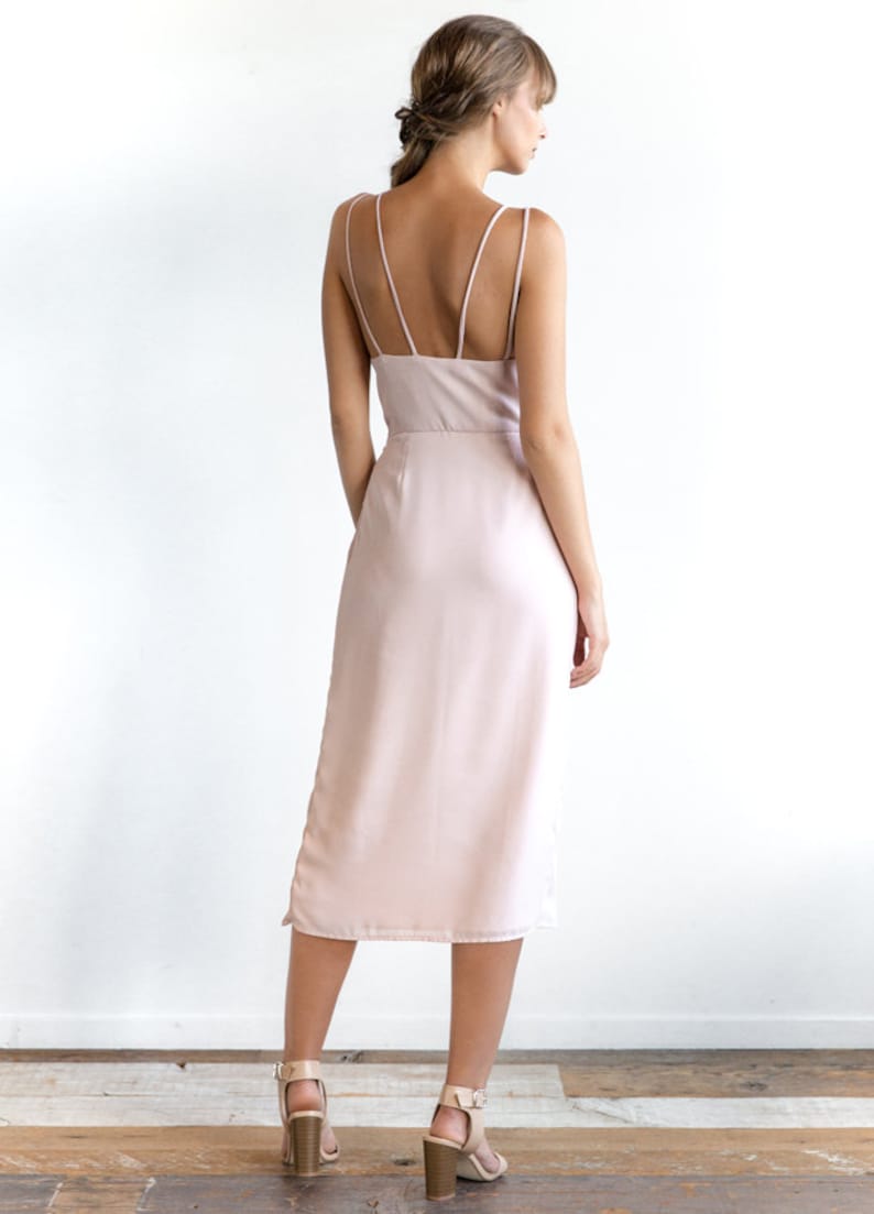 May include: A light pink midi dress with thin spaghetti straps and a simple, elegant design. The dress has a straight silhouette and a slightly longer length in the back.
