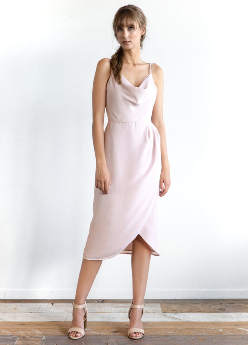 May include: A light pink midi dress with a cowl neck and thin straps. The dress has a wrap-style skirt and a fitted bodice.