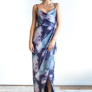 May include: A long, blue and purple floral print maxi dress with a cowl neck and a side slit. The dress has thin straps and a fitted bodice.
