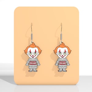 May include: A pair of dangle earrings featuring a cartoon clown design. The earrings have orange hair, white face, and a gray and red outfit. The earrings are displayed on a peach-colored background.