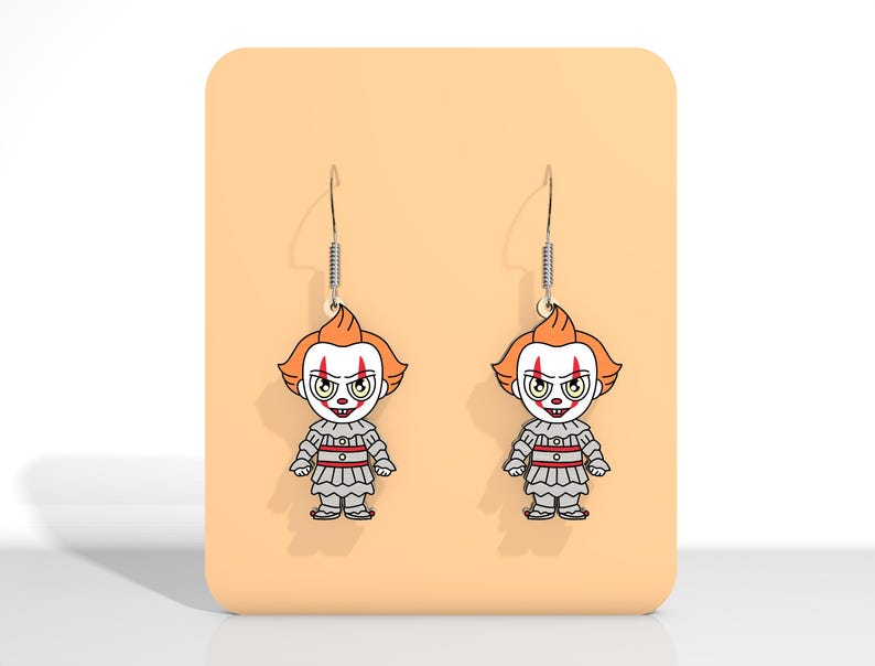 May include: A pair of dangle earrings featuring a cartoon clown design. The earrings have orange hair, white face, and a gray and red outfit. The earrings are displayed on a peach-colored background.