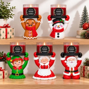 May include: Five holiday-themed candle holders shaped like a gingerbread person, snowman, elf, Mrs. Claus, and Santa Claus, each holding a lit White Barn Mahogany Teakwood candle. The figures are colorful, festive, and arranged on a wooden shelf.