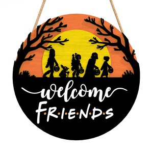 Pu&ograve; includere: Cartello rotondo in legno di Halloween con una silhouette nera di bambini che fanno dolcetto o scherzetto contro un tramonto arancione e giallo. Il cartello recita "welcome FRIENDS" in corsivo bianco e lettere maiuscole, con una corda di iuta per appenderlo.