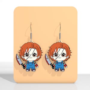 May include: A pair of dangle earrings featuring a cartoon Chucky doll design. Each earring depicts Chucky with red hair, blue overalls, and a knife. The earrings hang from silver-colored fish hooks.