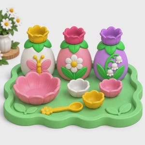 May include: A set of three egg-shaped containers with flower-shaped lids, three small bowls, and a yellow spoon on a green scalloped tray. The containers feature a butterfly, a daisy, and lily of the valley designs in pink, white, and purple colours.