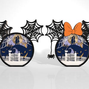 K&ouml;nnte beinhalten: Zwei Halloween-Dekorationen in Form von M&auml;useohren. Eine hat ein schwarzes Spinnennetz-Design und die andere eine orangefarbene Schleife. Jede zeigt eine mehrschichtige Szene mit Vollmond, Silhouetten und einer gruseligen Landschaft.