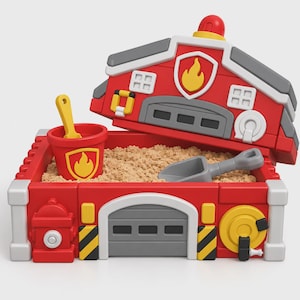 May include: A red and gray plastic fire station-themed sandbox filled with tan kinetic sand. The set includes a red bucket with a yellow flame emblem, a yellow shovel, and a gray scoop. The structure features a hinged lid with a red siren.