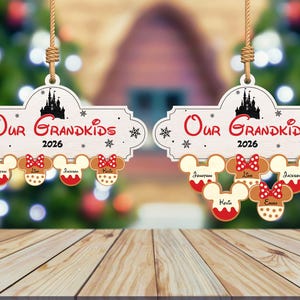 May include: Wooden Christmas ornament with the text "Our Grandkids 2026" and a silhouette of a castle. Below are gingerbread Mickey Mouse heads with names. The ornament hangs from a jute string.