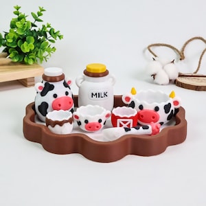 Puede incluir: Un juego de té en miniatura con temática de vaca sobre una bandeja marrón festoneada, que incluye una tetera con forma de vaca, una jarra de leche blanca con la etiqueta MILK, dos tazas pequeñas con estampado de vaca, una taza roja y una cuchara.