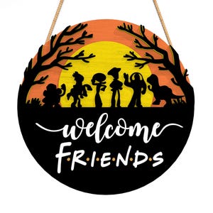 Pu&ograve; includere: Un cartello rotondo in legno con sagome nere di personaggi dei cartoni animati su uno sfondo di tramonto arancione e giallo. Il cartello recita "welcome FRIENDS" in scrittura bianca. Una corda marrone &egrave; attaccata per appenderlo.