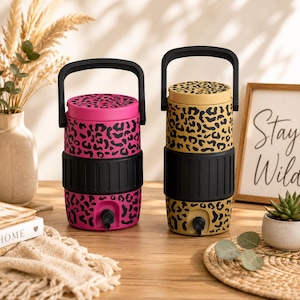 May include: Two cylindrical beverage dispensers with black handles and spigots. One dispenser is bright pink with a black leopard print, and the other is tan with a black leopard print. Both feature a wide black ribbed band around the centre.