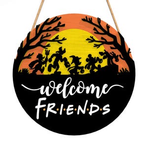 Pu&ograve; includere: Cartello rotondo in legno di Halloween con una silhouette nera di personaggi dei cartoni animati in costume contro un tramonto arancione e giallo. Le parole "welcome friends" sono scritte in bianco. Appeso con una corda naturale.