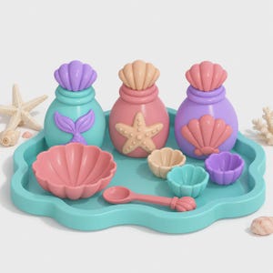 Puede incluir: Un juego de tres recipientes redondeados con tapas en forma de concha, un cuenco grande, tres cuencos pequeños y una cuchara, todo dispuesto sobre una bandeja festoneada de color turquesa en tonos pastel variados.