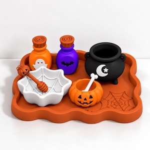 Puede incluir: Un set temático de Halloween sobre una bandeja naranja que incluye un cuenco blanco de telaraña con un cucharón de miel de calabaza, una botella naranja de fantasma, una botella morada de murciélago, un caldero negro y un cuenco de calabaza.