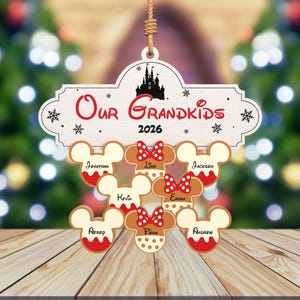 May include: A wooden Christmas ornament with the words "Our Grandkids 2026" and a silhouette of a castle. Below are gingerbread Mickey Mouse heads with names. The ornament is hanging in front of a blurred Christmas tree.