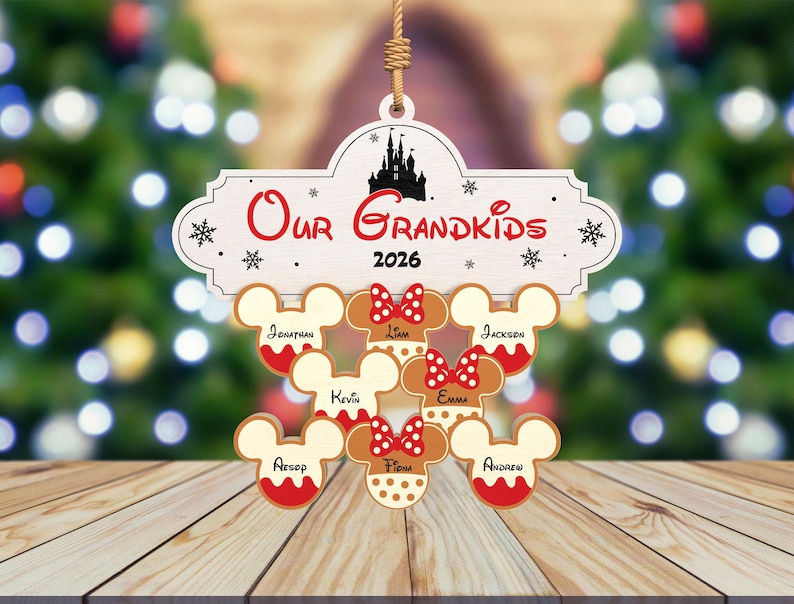 May include: A wooden Christmas ornament with the words "Our Grandkids 2026" and a silhouette of a castle. Below are gingerbread Mickey Mouse heads with names. The ornament is hanging in front of a blurred Christmas tree.