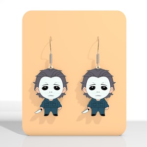 May include: A pair of dangle earrings featuring a cartoon depiction of a character with a white mask, gray hair, and a blue jumpsuit, holding a knife. The earrings hang from silver-colored hooks and are displayed on a peach-colored background.