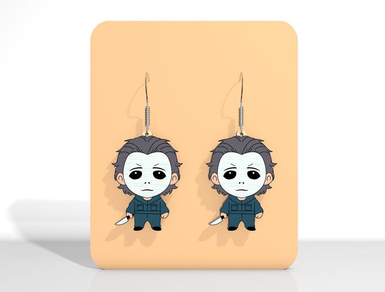 May include: A pair of dangle earrings featuring a cartoon depiction of a character with a white mask, gray hair, and a blue jumpsuit, holding a knife. The earrings hang from silver-colored hooks and are displayed on a peach-colored background.