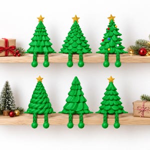 May include: Six green plastic Christmas tree figurines with articulated dangling legs and gold star toppers. Each tree features a unique textured design, with one tree decorated with small colourful spheres, all arranged on two wooden shelves.