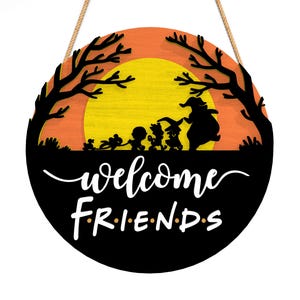 Pu&ograve; includere: Cartello di benvenuto rotondo in legno a tema Halloween. Il cartello presenta sagome nere di personaggi e alberi su uno sfondo arancione e giallo. Le parole "welcome FRIENDS" sono scritte in bianco.