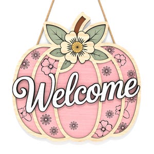 May include: A wooden pumpkin-shaped welcome sign with a pink center and floral accents. The word "Welcome" is written in white script across the front. The sign is decorated with a flower and leaves at the top and is suspended by a natural rope.