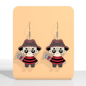 May include: A pair of novelty earrings featuring a cartoon character with a brown hat, red and black striped sweater, and metal clawed glove. The earrings are displayed on a peach-colored card.