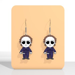 May include: A pair of Halloween-themed dangle earrings. Each earring features a cartoon illustration of a character in a blue jumpsuit, white mask, and holding a knife. The earrings hang from silver-colored fish hook ear wires.