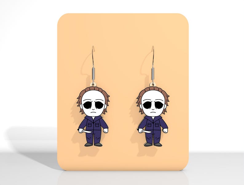 May include: A pair of Halloween-themed dangle earrings. Each earring features a cartoon illustration of a character in a blue jumpsuit, white mask, and holding a knife. The earrings hang from silver-colored fish hook ear wires.