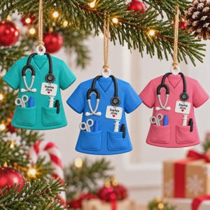 May include: Three hanging ornaments shaped like medical scrub tops in teal, blue, and pink. Each features a black stethoscope, a white ID badge with the text Sophia 2026 and a red heart, and pockets containing scissors, a pen, and a syringe.