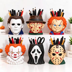 Op de afbeelding: Een set van zes bureau-organizers met een horror-thema in de vorm van iconische filmkarakterhoofden, waaronder Michael Myers, Freddy Krueger, Chucky, Pennywise, Ghostface en Jason Voorhees, elk gevuld met diverse pennen, potloden en kantoorbenodigdheden.