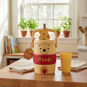 May include: A yellow beverage dispenser featuring a Winnie the Pooh design with a red band labeled Pooh, a top handle, a spigot, and a small honey pot tag. The dispenser is accompanied by a matching yellow plastic cup, creating a cohesive set.
