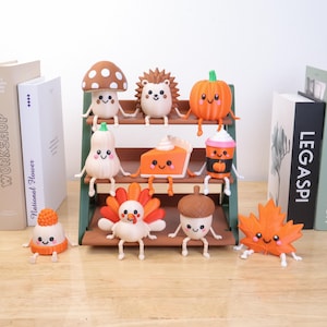 May include: A three-tiered green display shelf holds nine small, cartoon-style figurines with smiling faces. The collection includes a mushroom, hedgehog, pumpkin, gourd, slice of pie, coffee cup, turkey, acorn, and a maple leaf figurine.