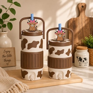 May include: Two cylindrical tumblers with brown and white cow print patterns, brown lids with handles, and light blue straws. Each lid features a small cartoon highland cow topper with a pink bow. The tumblers have a brown ribbed grip section.